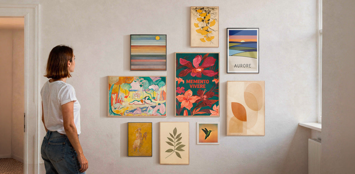 A woman stands in a doorway looking at a gallery wall of framed art prints in a bright, modern room. The collection includes various colorful abstract, floral, and landscape designs in different frame styles.